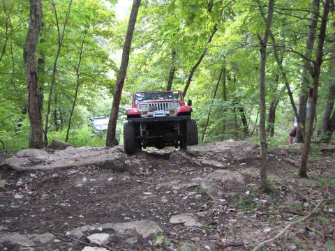 2005 Jeep Wrangler - A red Jeep Wrangler drives through a wooded area with large rocks on the path.