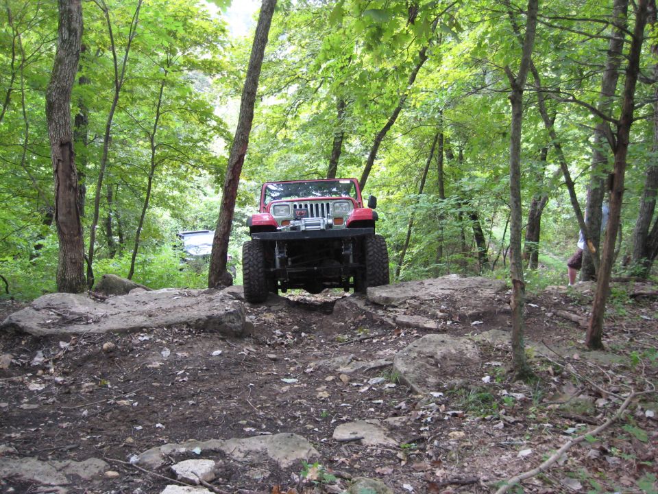 Red Jeep Wrangler with a winch on the front bumper navigates a rocky, wooded trail