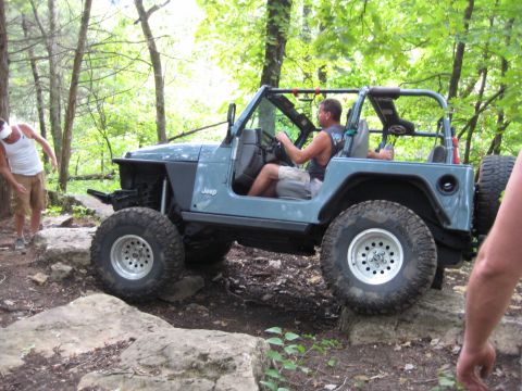 2005 Jeep Wrangler - A blue Jeep Wrangler is shown off-roading on a rocky trail with trees in the background.