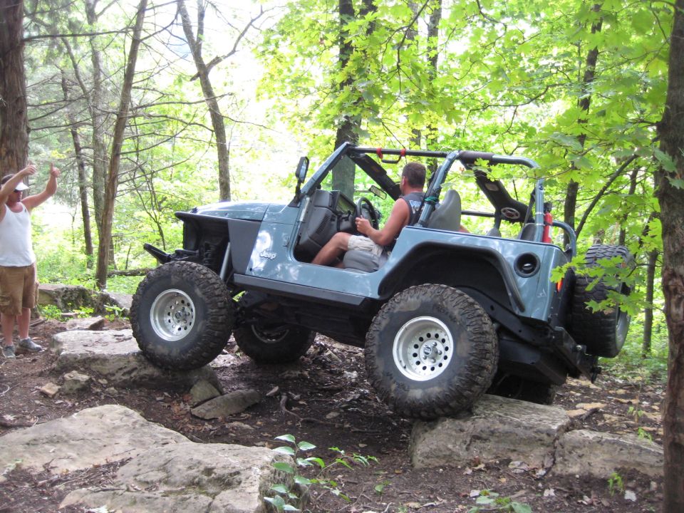 Blue 2005 Jeep Wrangler driving over rocks on a wooded trail. Driver is visible. Another person stands nearby