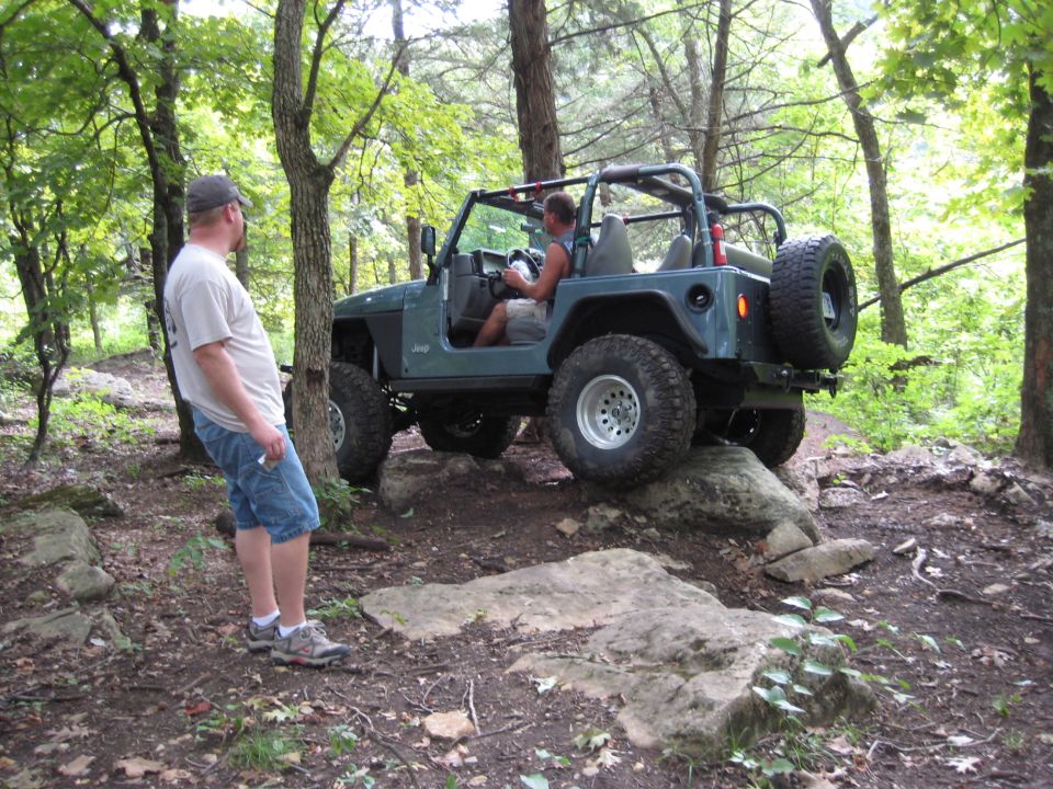 Guy watches a green Jeep Wrangler drive over rocks on a wooded trail. The Jeep has a roll cage and a spare tire on the back