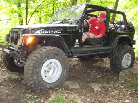 2005 Jeep Wrangler - Low side view of a lifted black Jeep Wrangler Rubicon with doors and top removed, flexing over rocks on a wooded dirt trail. It rides on ov…