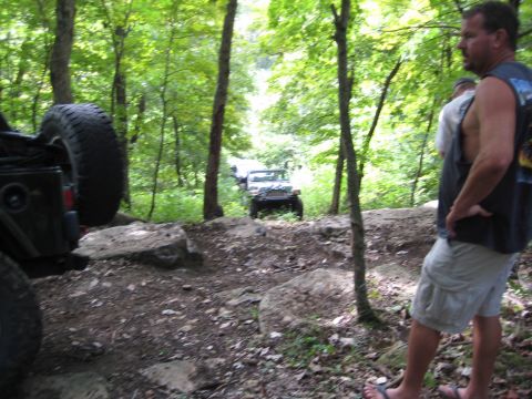 2005 Jeep Wrangler - A dark green Jeep with an oversized swing-out spare tire sits tilted on a rocky wooded trail at left while a white Jeep with a winch bumper…