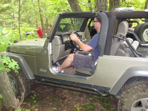 2005 Jeep Wrangler - A man drives a 2005 Jeep Wrangler through a wooded area, with another vehicle visible in the background.