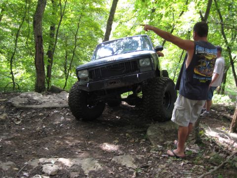2005 Jeep Wrangler - A man points towards a lifted Jeep Cherokee XJ navigating a wooded, rocky path.