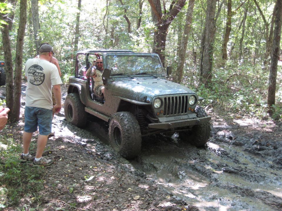 2005 Jeep Wrangler navigating a muddy trail in a wooded area. Another Jeep is partially visible in the background