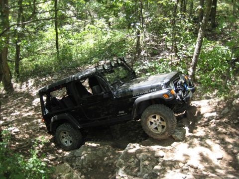 2005 Jeep Wrangler - A black Jeep Wrangler Rubicon navigates a challenging rocky terrain on a wooded trail.