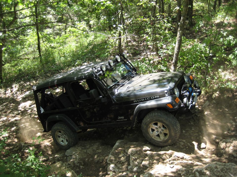 Black 2005 Jeep Wrangler Rubicon driving over rocks on a wooded trail. Light bar on roof