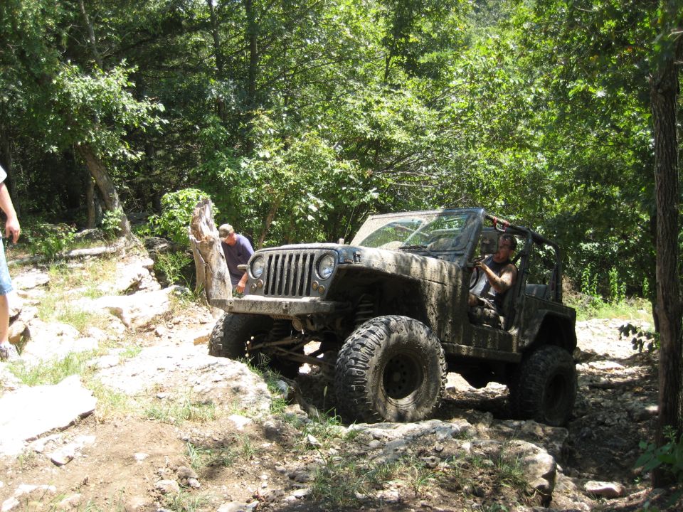Mud-covered 2005 Jeep Wrangler driving on a rocky, wooded trail