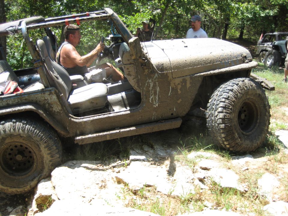 Mud-covered 2005 Jeep Wrangler, 3/4 front view, driving over rocks with driver inside