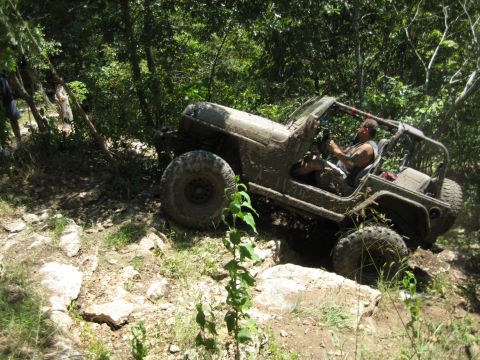 2005 Jeep Wrangler - A muddy Jeep Wrangler TJ navigates a challenging off-road course with large rocks and dense foliage.