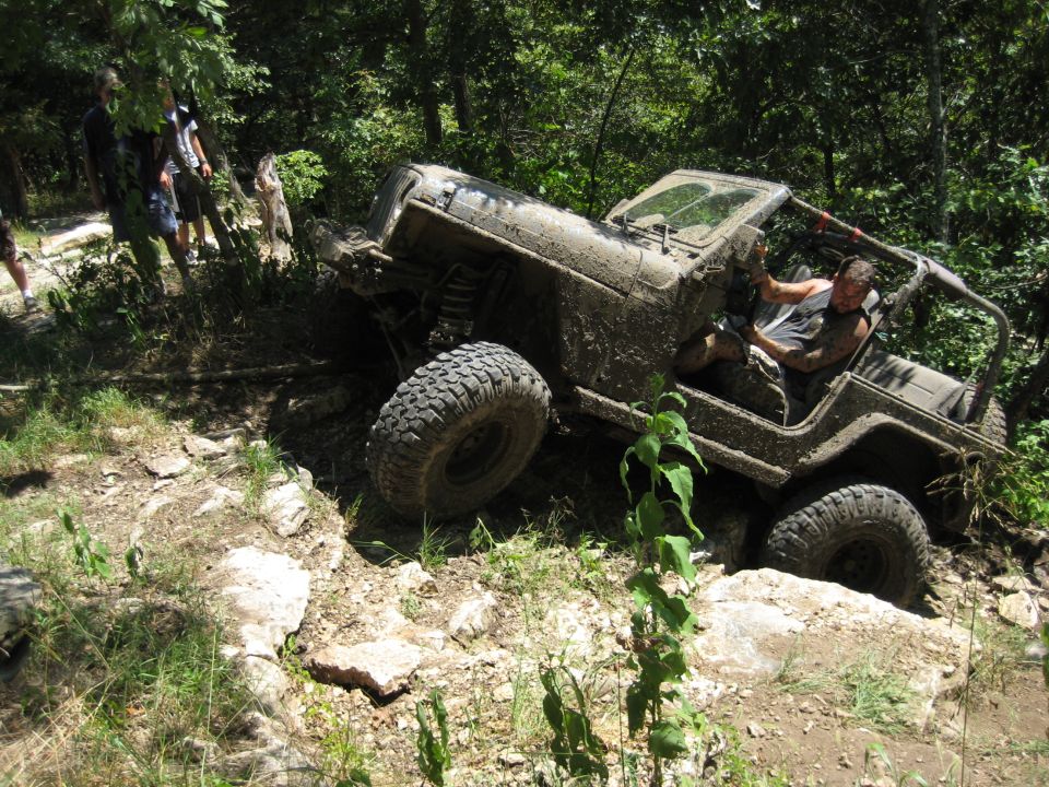 Muddy Jeep Wrangler struggling on a rocky, wooded trail with a driver inside