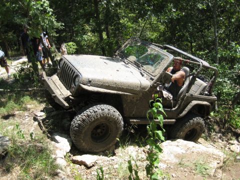 2005 Jeep Wrangler - A muddy Jeep Wrangler navigates a challenging off-road course with trees and rocks surrounding it.