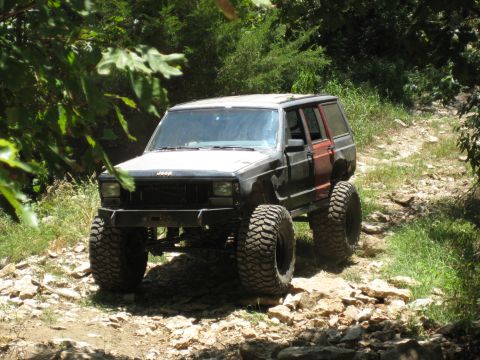 2005 Jeep Wrangler - This modified Jeep Cherokee XJ is navigating a rough, rocky off-road path surrounded by trees.