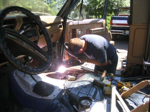 1986 Ford Bronco - A person welds inside a vehicle's interior while tools lie nearby; another truck is parked in the background.
