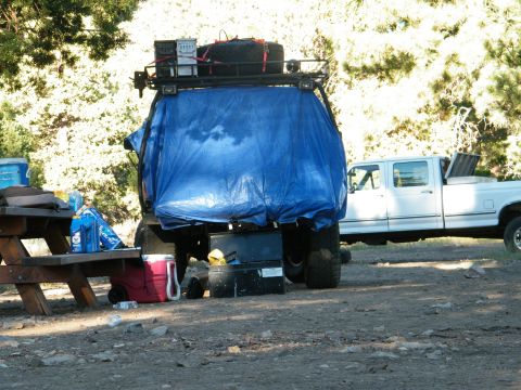 1982 Ford Bronco - An off-road vehicle, mostly covered by a blue tarp, is parked next to a white pickup truck.