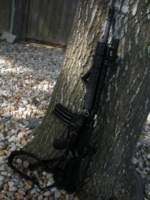 1996 Ford Bronco - An assault rifle is propped against a tree, with a wooden fence and gravel in the background.