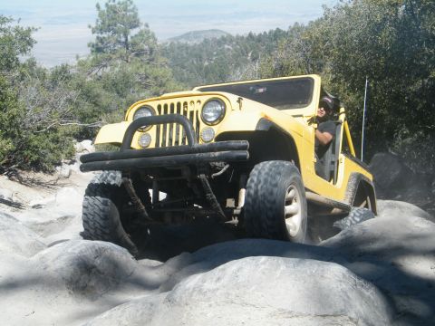 1982 Ford Bronco - A yellow Jeep Wrangler tackles a challenging rocky trail in a mountainous, wooded area.