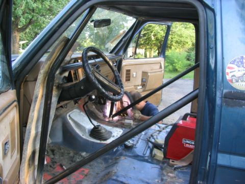 1986 Ford Bronco - A person welds a roll cage inside a truck's interior, with the driver's side door open.