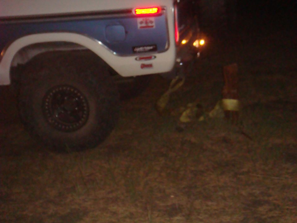 Rear quarter of a 1978 Ford Bronco with a blue stripe and a spare tire mounted on the rear door. Straps are on the ground