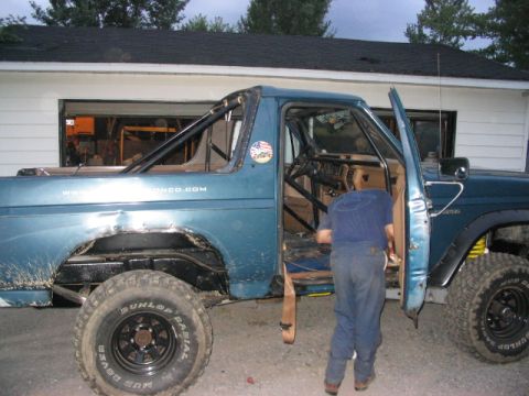 1986 Ford Bronco - A blue SUV, possibly a Ford Bronco, is shown with an open door and a visible roll cage, with a website address on its side.
