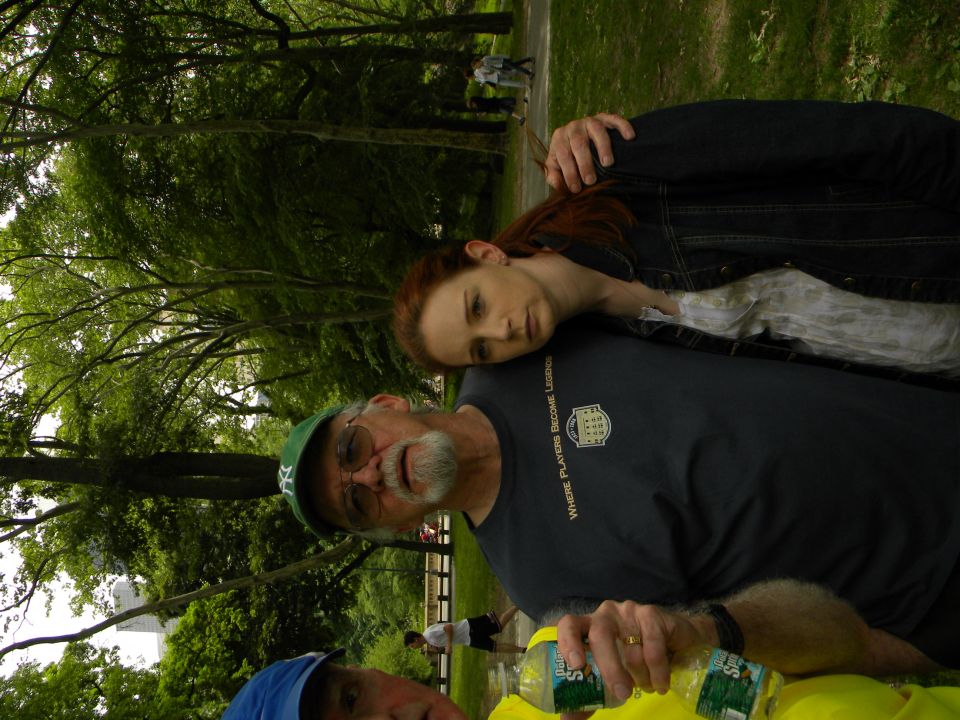 People in a park with trees and grass, one man wears a green cap, another a blue cap, and a woman rests her head on a man's shoulder