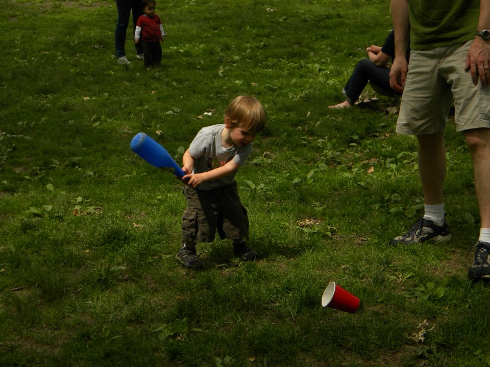 Child with blue plastic bat swings at red cup on grass, with adults and another child in background