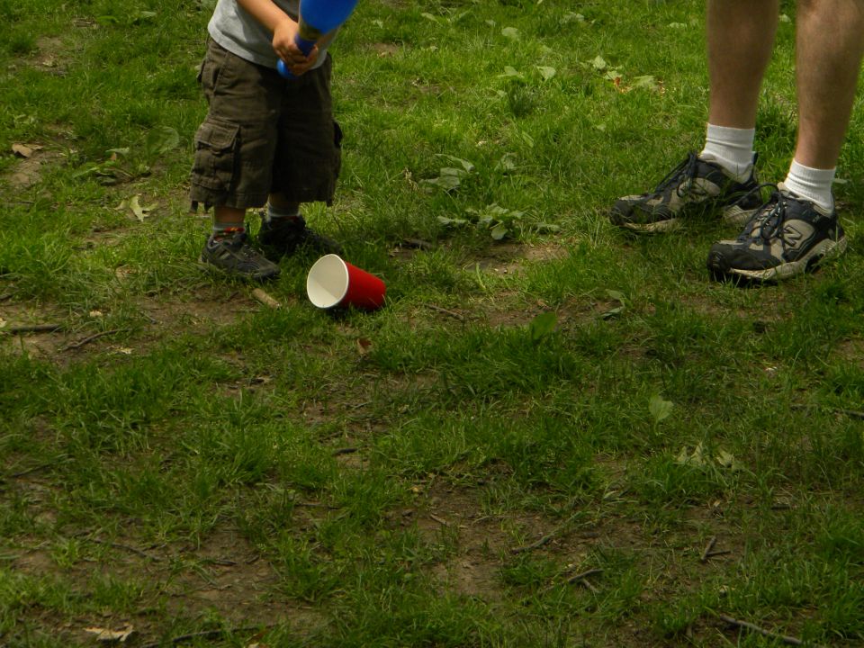 Child in cargo shorts and adult in sneakers stand on grass near a red cup