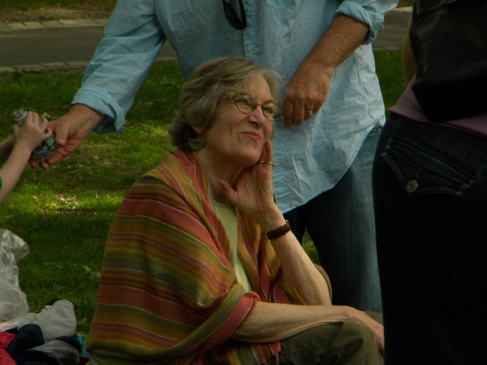 Woman with glasses and a striped shawl sits outdoors, with people behind her