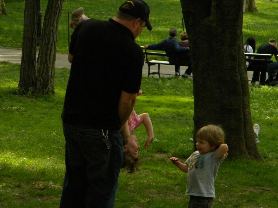 Man holding a child upside down in a park, another child stands nearby