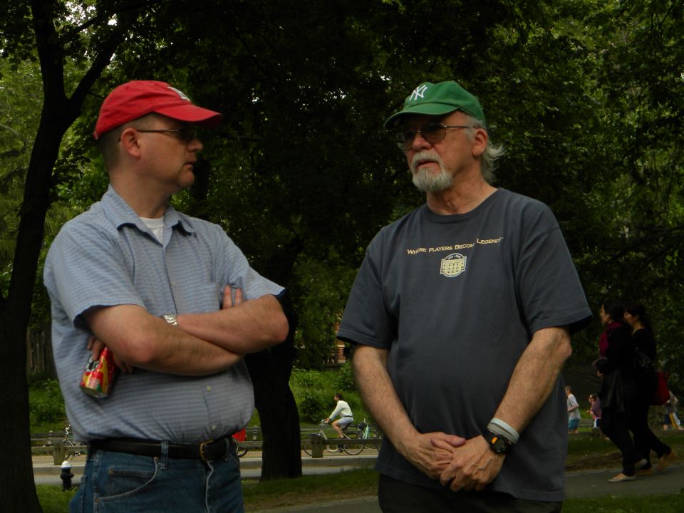 Two men talking in a park, one wearing a red cap, the other a green cap