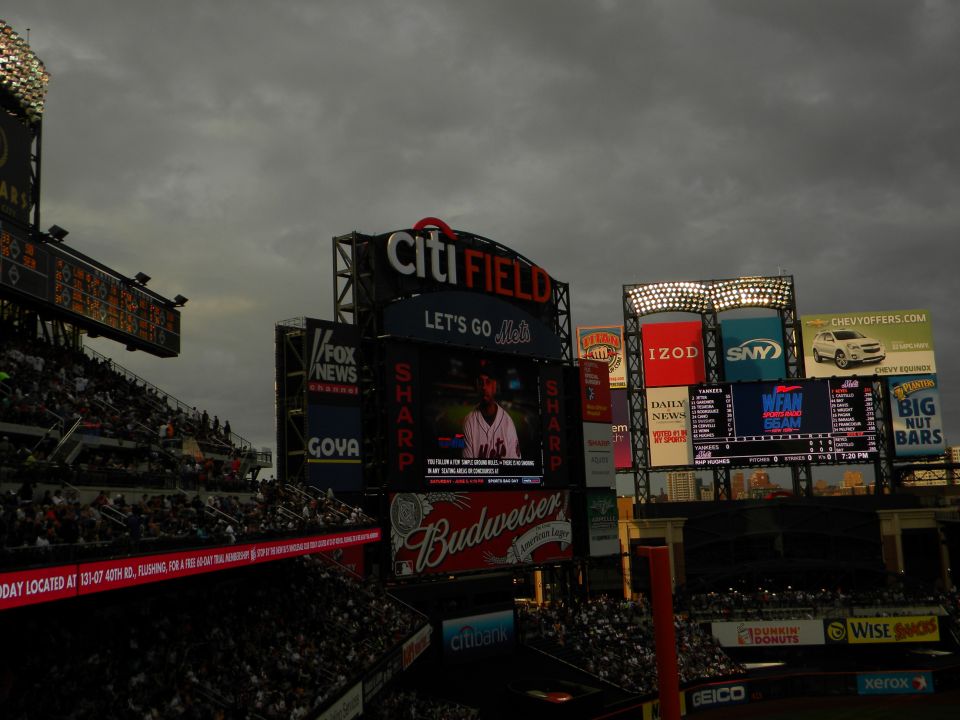 Citi Field stadium exterior with multiple large screens displaying ads and game information under a cloudy sky
