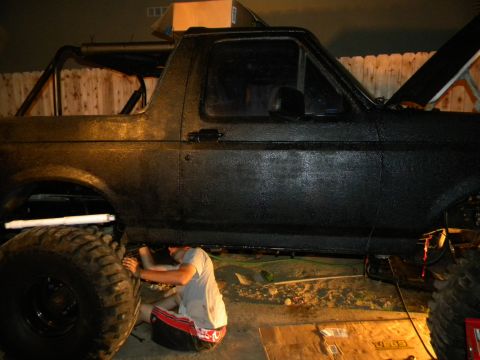 1992 Ford Bronco - A person works on the underside of a black Ford Bronco with oversized tires, parked outdoors at night.
