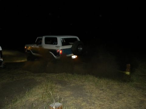 1992 Ford Bronco - White Ford Bronco driving on a dirt road at night, kicking up dust. Another vehicle is partially visible.