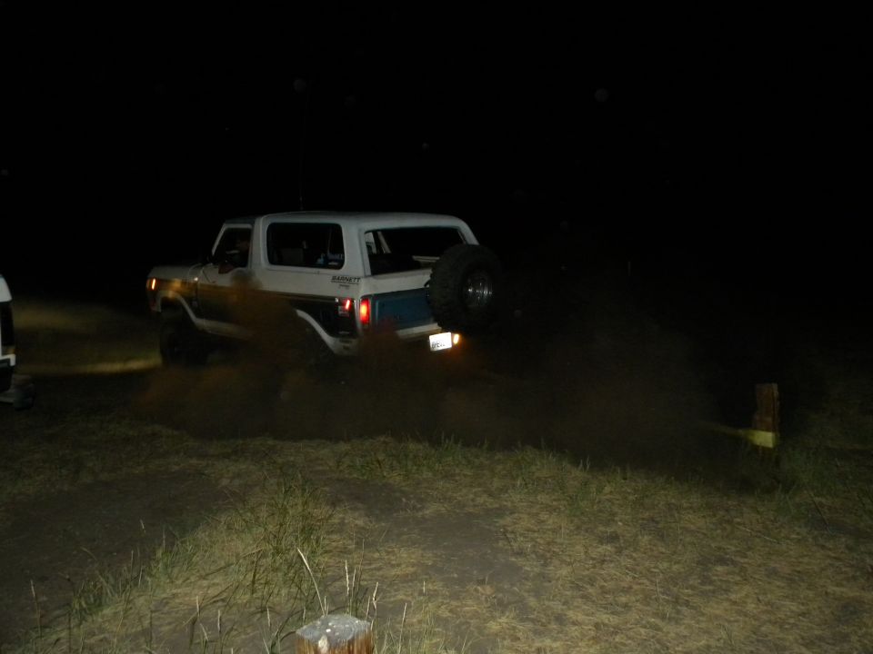 White Ford Bronco with spare tire on back driving on dirt road at night, kicking up dust. Another vehicle is partially visible on the left