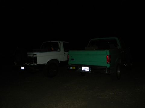 1992 Ford Bronco - Two trucks, a white 1992 Ford Bronco and a green pickup, are parked side-by-side at night.