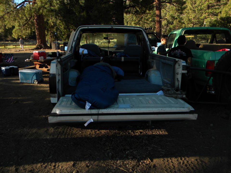 Pickup truck bed with a blue sleeping bag and camping gear. Another green truck is parked nearby