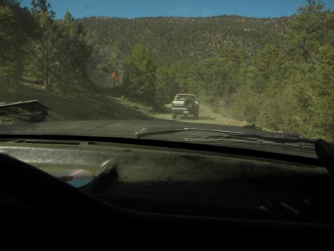 1992 Ford Bronco - A white pickup truck travels down a dusty dirt road surrounded by trees, with another vehicle visible ahead.