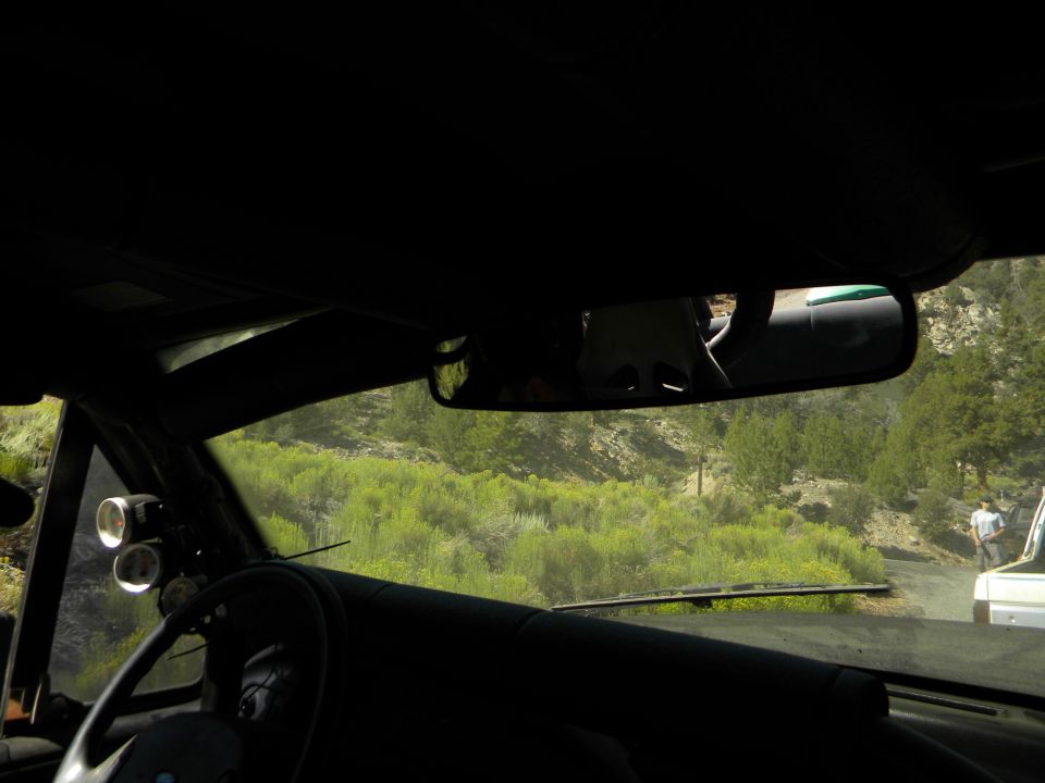 Interior view from a vehicle, looking out the windshield at a dirt road and green hills. Another vehicle is visible on the road