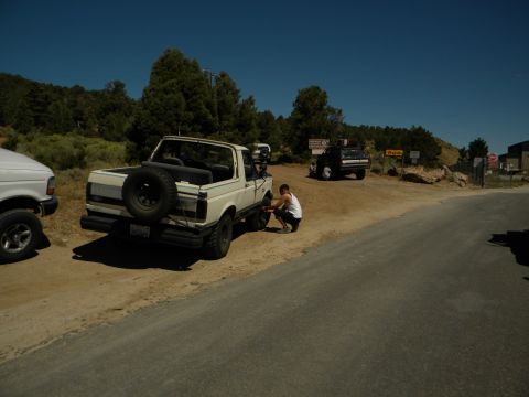 1992 Ford Bronco - A person works on the front tire of a white Ford Bronco parked on a dirt road with other vehicles nearby.