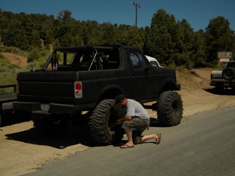 1992 Ford Bronco - Man works on a lifted black Ford Bronco with oversized tires. Another vehicle is visible in the background.