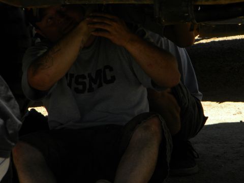 1992 Ford Bronco - Two individuals are crouched down, appearing to work on the underside of a vehicle in a sunny outdoor setting.