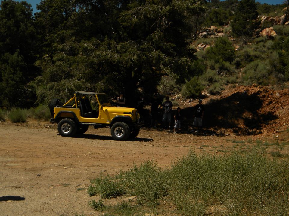 Yellow Jeep CJ with a roll cage and soft top parked on a dirt road with trees and hills in the background