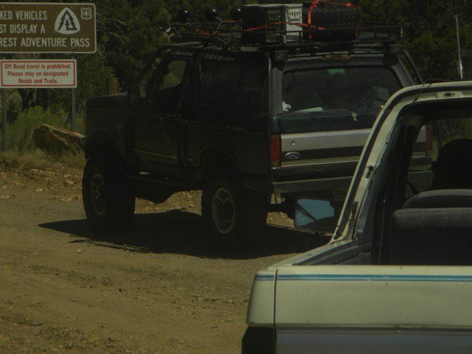1992 Ford Bronco with roof rack parked on a dirt road, another vehicle behind it. Signage visible