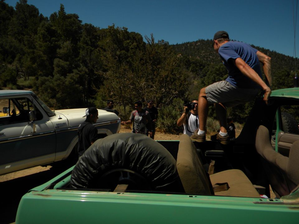 Green truck bed with spare tire, white and blue truck beside it, people in a wooded area