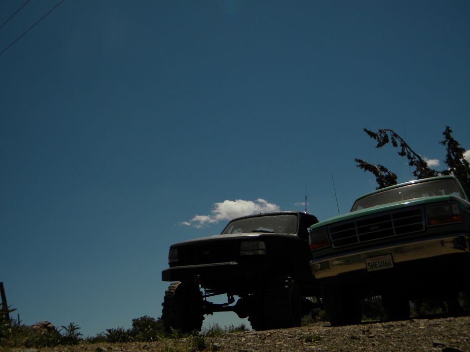 Black 1992 Ford Bronco and a teal 1990s Ford F-Series truck parked on a dirt incline under a blue sky