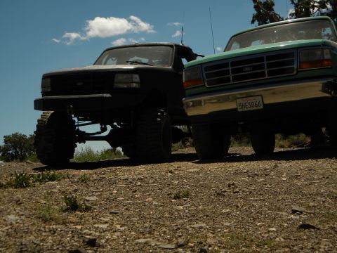 1992 Ford Bronco - A black 1992 Ford Bronco with a lifted suspension sits next to a teal truck under a blue sky.