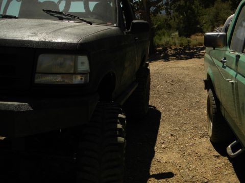 1992 Ford Bronco - Two lifted SUVs, one a 1992 Ford Bronco, parked side-by-side on a dirt road.