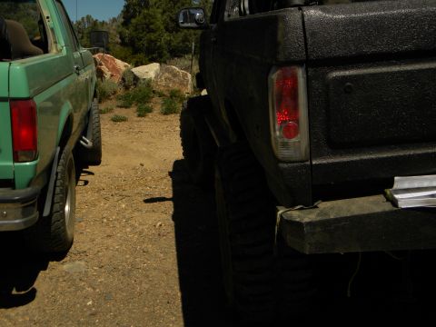 1992 Ford Bronco - Two lifted Ford Broncos parked on a dirt lot with rocks and trees in the background.