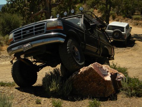 1992 Ford Bronco - A black Ford truck navigates a rocky trail, with a white SUV visible on the dirt road behind it.